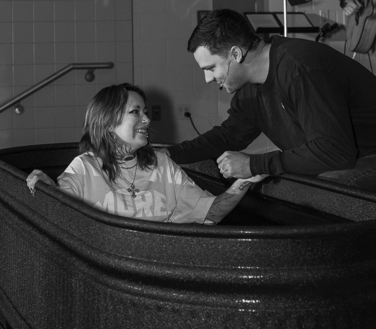 A woman standing in the baptism tank, smiling up at the pastor as he leans in and holds her arm