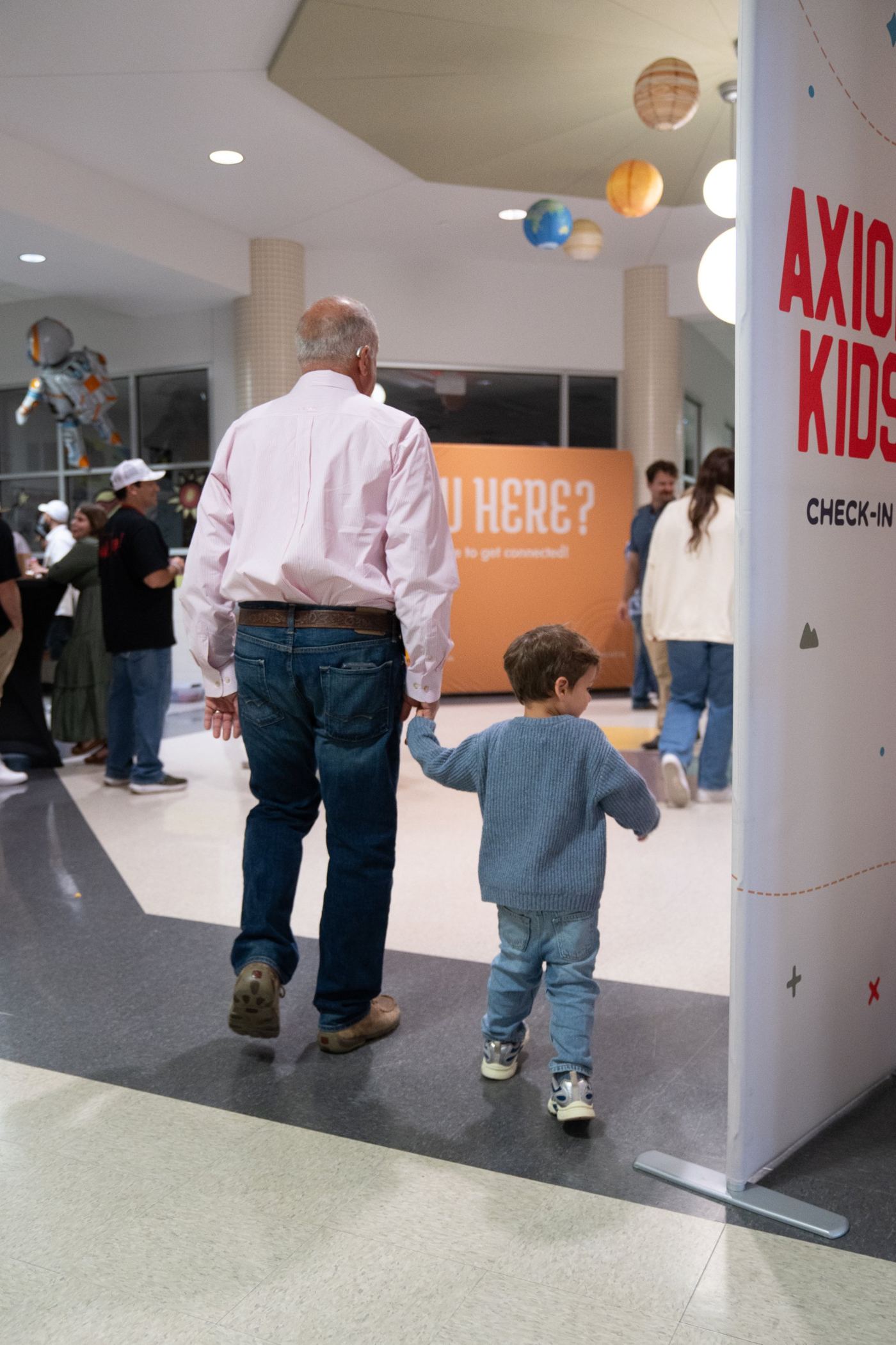 A grandfather holding his grandson’s hand as they walk past the Axiom Kids check-in sign