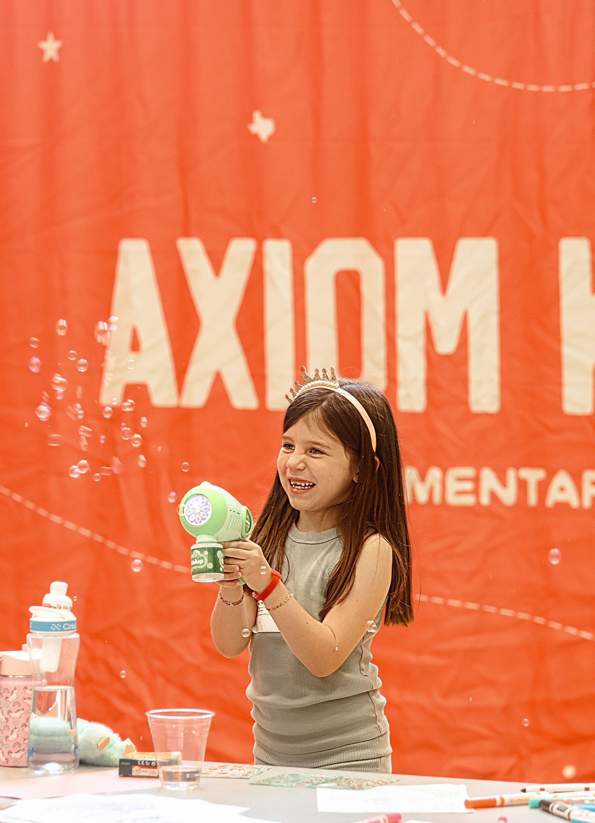A girl in a tiara laughing as she fires a bubble gun in front of the Axiom Kids banner