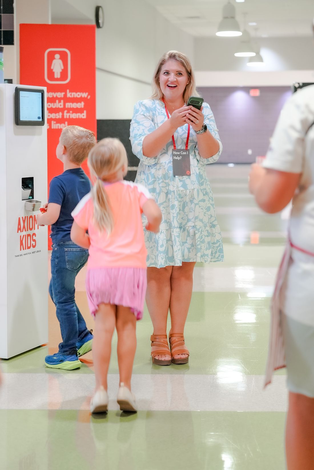 A volunteer wearing a How Can I Help lanyard greets two kids at the Axiom Kids check-in kiosk