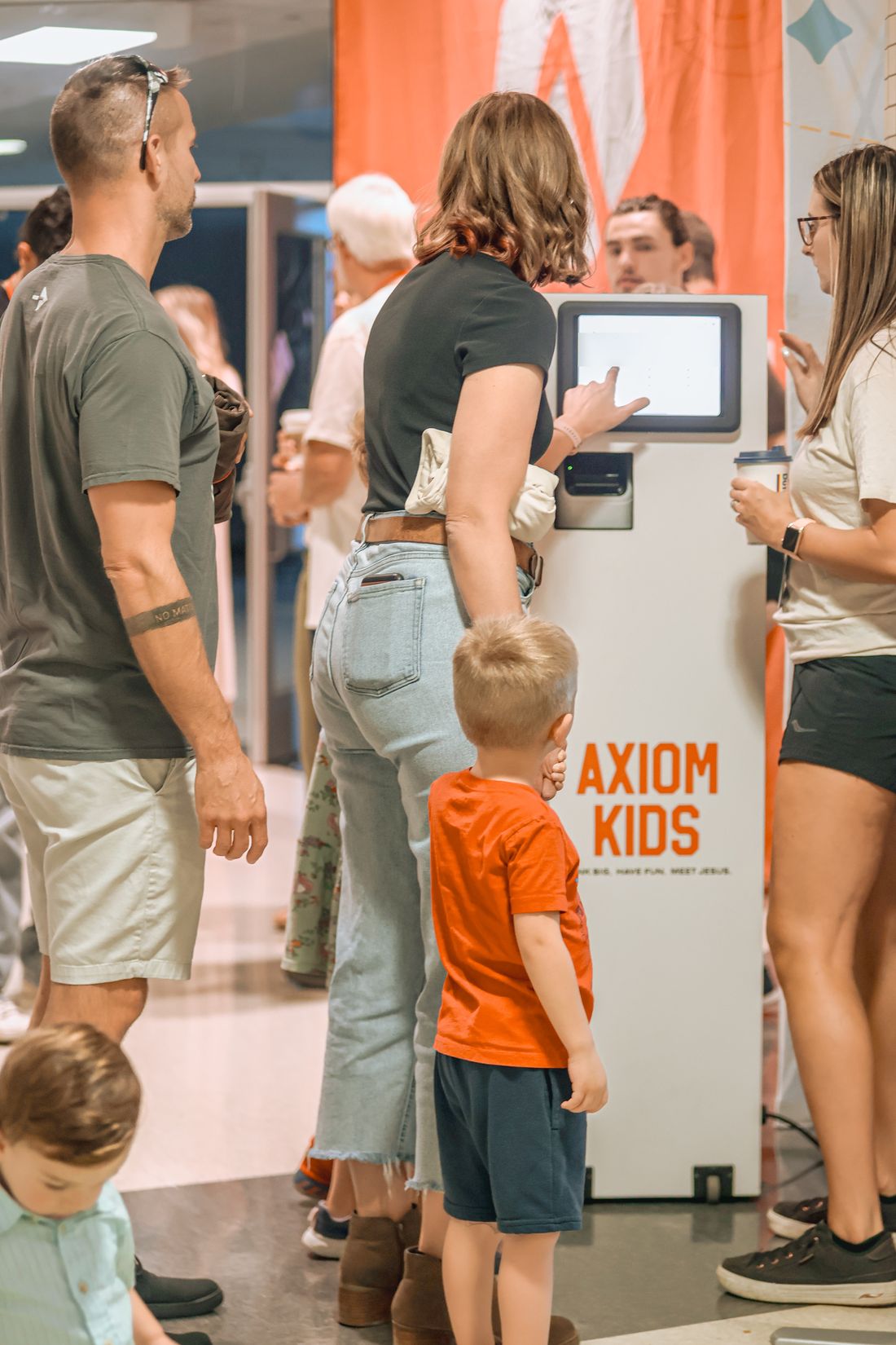 A mom typing her phone number into the Axiom Kids check-in kiosk while her son waits beside her