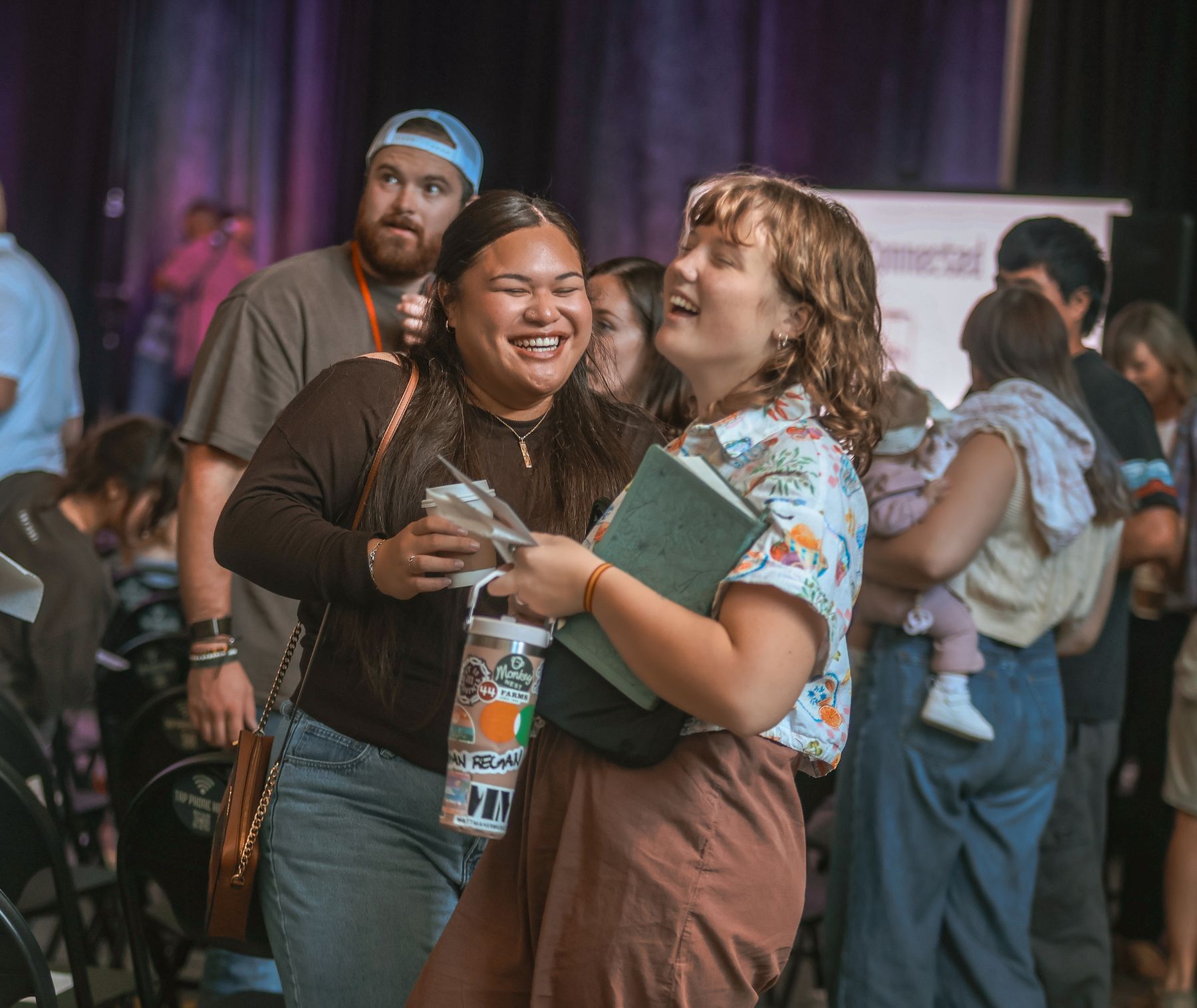 Two women laughing together after a Sunday service, one holding a coffee
