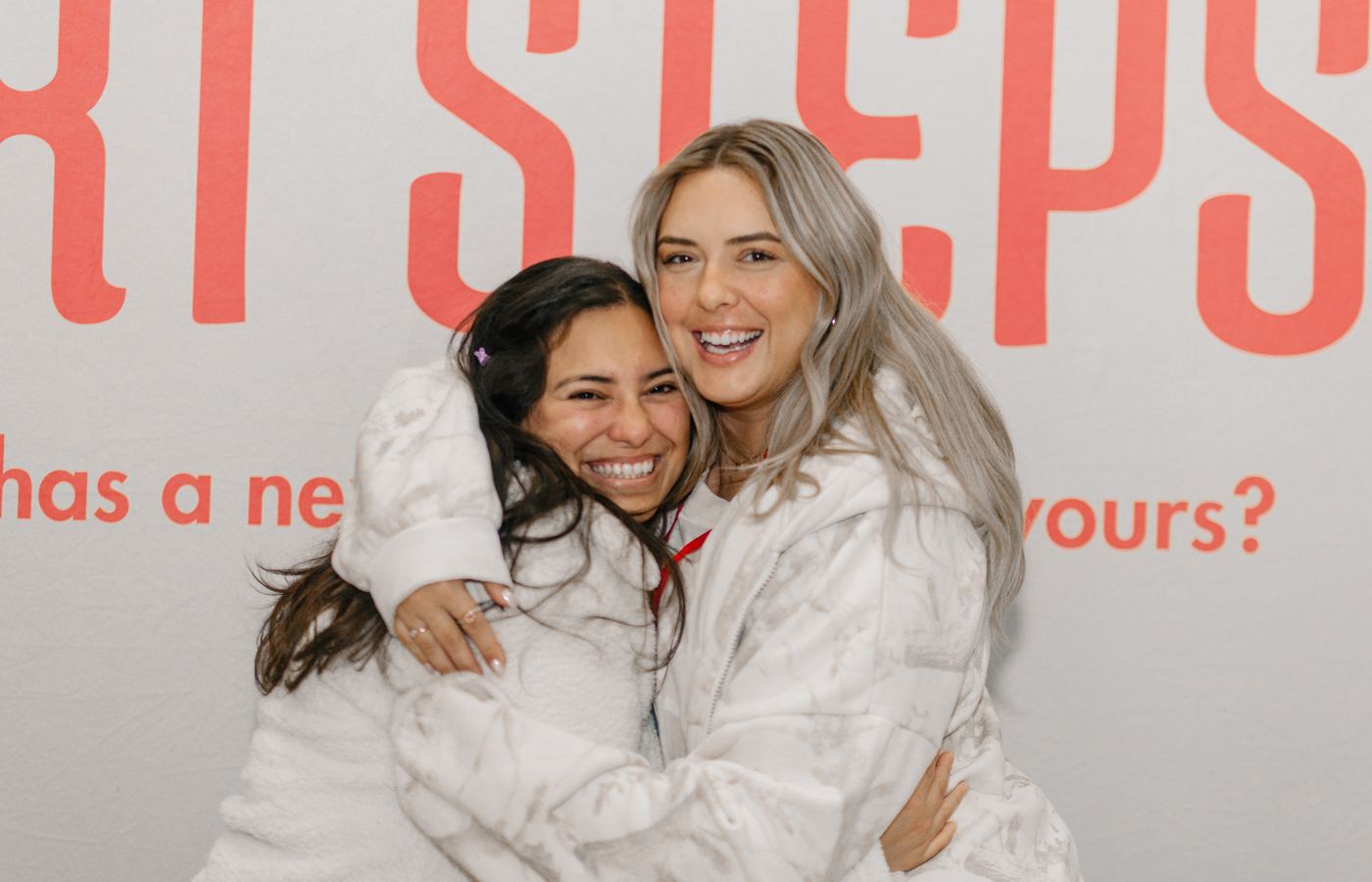Two women in white jackets hugging and laughing in front of the Next Steps banner