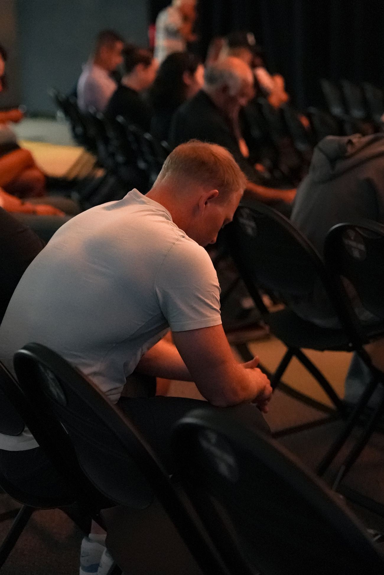 A man in a white T-shirt leaning forward in his chair, head bowed in prayer during a Sunday service