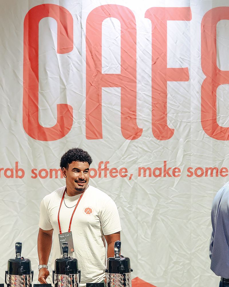 A volunteer smiling behind the coffee pots under the Cafe banner