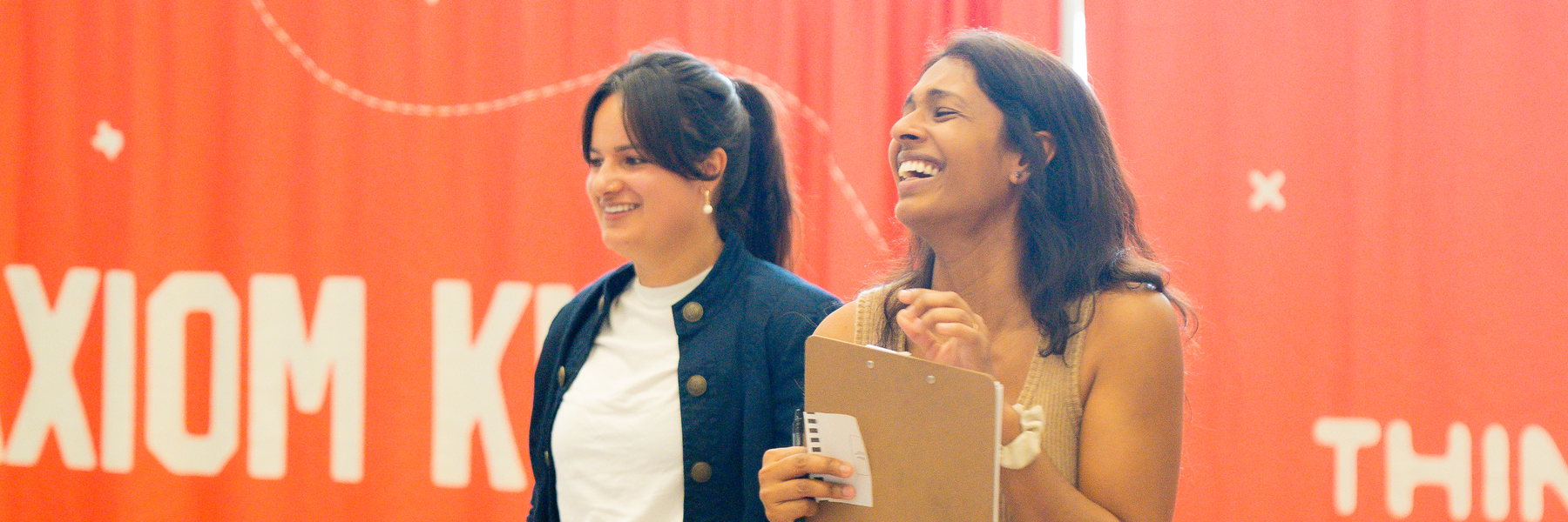 Two Axiom Kids volunteers laughing in front of the red Axiom Kids banner