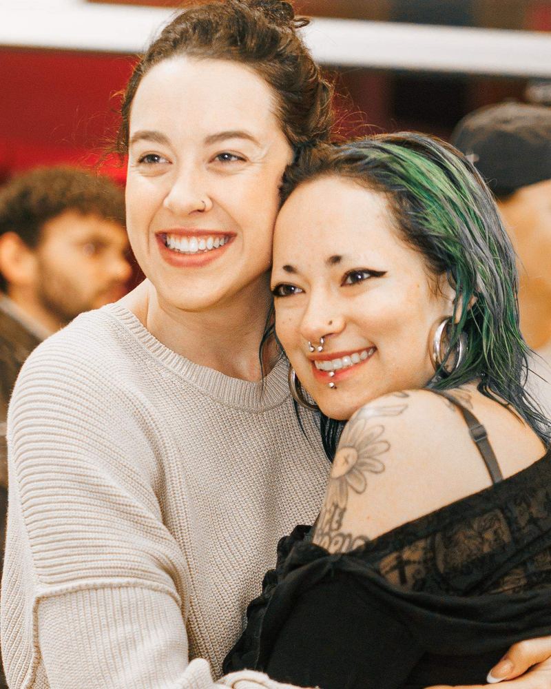 Two women hugging, one with tattoos and green hair, both smiling